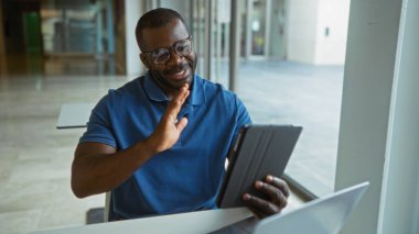 Ofisteki genç adam video görüşmesi sırasında tablete el sallıyor ve modern iç mekanlı profesyonel bir çalışma ortamı sergiliyor.