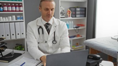 Klinikte çalışan orta yaşlı, beyaz önlüklü, steteskoplu beyaz bir beyaz adam. Tıbbi malzemeler arasında dizüstü bilgisayara yoğunlaşmış..