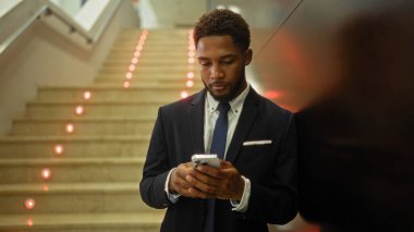 Bir ofis binasının içinde duran genç adam, merdivenlerin yanındaki duvara yaslanırken telefonuna bakıyor.
