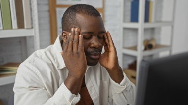 Ofisteki genç adam gözleri kapalı stresli görünüyor, modern bir iş yerinde elleriyle şakaklarını bastırıyor..