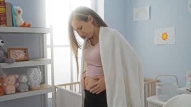 Sıcak bir yatak odasında duran kadın karnına dokunuyor. Bebek oyuncakları ve beşikle çevrili. Sıcak bir ev ortamı yaratıyor..