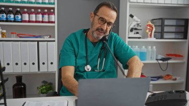 Hastane kliniğinde çalışan olgun bir adam telefonla konuşuyor. Steteskop altında yeşil önlük, mikroskop ve düzenli iş yerinde görülebilen raflar var.