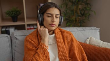 Kanepede dinlenen, kulaklık takan, gözleri turuncu battaniyeye sarılı rahat oturma odasında bitki ve kitaplarla müzik dinleyen bir kadın.