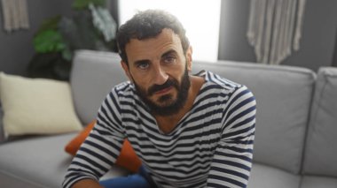 Salonda oturan sakallı yakışıklı, çizgili tişörtlü ve günlük atmosferli bir adam.