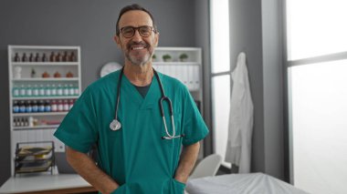 Ameliyat önlüğü ve steteskop giyen olgun İspanyol bir adam hastane kliniğinde güvenle gülümsüyor ve tıbbi bir ortamda profesyonel ve şefkatli bir atmosfer yaratıyor.