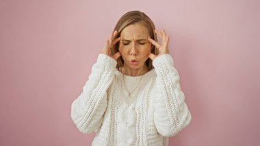 Pembe arka planda strese girmiş bir kadın, beyaz bir kazak giyerken şakaklarına dokunuyor, gerginlik ve hayal kırıklığını iletiyor..