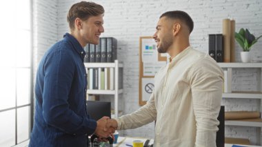 Işıl ışıl bir ofiste el sıkışan iki adam, arka planda ofis malzemeleriyle iş anlaşması ya da ortaklık anlamına geliyor, kapalı alanda profesyonel bir iş ortamı sergiliyorlar.