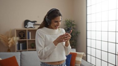 Kulaklıkla gülümseyen bir kadın modern oturma odasında akıllı telefon kullanarak doğal ışık ve şık dekorlarla rahat bir yaşam tarzı ve dijital bağlantı sağlıyor..