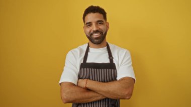 Kollarını kavuşturup, izole edilmiş sarı bir arka plana güvenen çekici İspanyol bir adam.