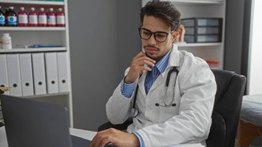 Klinikte laboratuvar önlüğü giyen bir adam laptopta çalışıyor ve arka planda tıbbi ekipmanlar var..