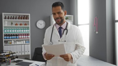 Genç İspanyol erkek doktor klinikte gülümsüyor. Elinde raflı belgeler ve arka planda profesyonel bir sağlık sigortasını gösteren bir saat var..