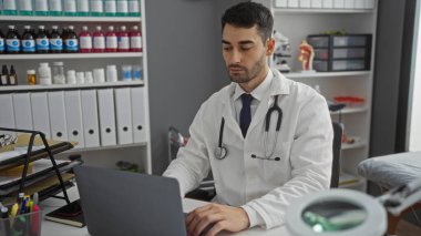 Klinikte çalışan Latin kökenli genç bir adam. Etrafı ilaç şişeleriyle çevrili dizüstü bilgisayar kullanıyor..