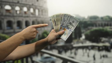 Roma Kolezyumu 'nun yakınında tutulan Amerikan dolarlarını gösteren bir el, şehir arka planındaki bir seyahat ve turizm bağlamında paranın altını çiziyor..