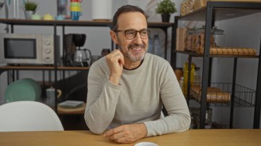 Kahve makinesi, tabak ve modern dekor ile çevrili bir ofis mutfağında gelişigüzel gülümseyen olgun İspanyol bir adam profesyonel ama rahat bir atmosfer sergiliyor..