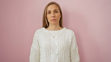 İzole edilmiş pembe arka planda duran beyaz kazaklı bir kadın. Stüdyo ortamında sakin, sarı saçlı ve düşünceli bir ifade takınıyor..