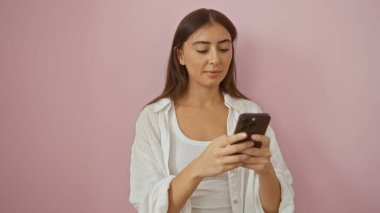 Esmer, zeki bir kadın pembe arka plan üzerinde akıllı telefon kullanıyor. Modern ve çekici İspanyol bir kadının günlük kıyafetlerini giyiyor.