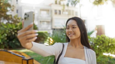 Şehir parkında, arka planda yeşillik ve şehir binalarıyla çevrili akıllı bir telefonla selfie çeken bir kadın, açık hava güzelliğinin ve modern şehir hayatının bir anını yakalıyor..