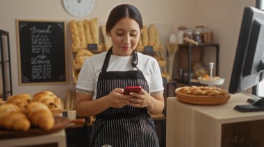 Fırında akıllı telefon kullanan genç bir kadın. Önlük takıyor ve arka planda raflar var.