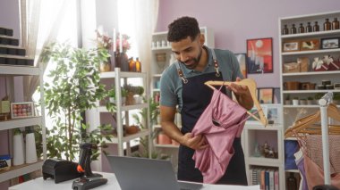 Ev dekorasyonu mağazasında genç bir adam elinde tarayıcıyla bir masanın arkasında dururken pembe bir giysiyi inceliyor. Etrafı çeşitli dekoratif eşya ve bitkilerle dolu raflarla çevrili.