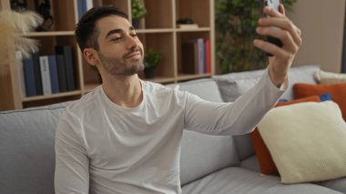 Genç bir adam rahat bir oturma odasında selfie çekiyor, modern bir ev dekorasyonu sergiliyor..