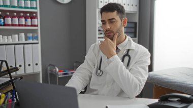 Hastane ofisinde steteskoplu labaratuvar önlüğü giyen genç İspanyol doktor gündüz vakti evde bilgisayarla uğraşmayı düşünüyor.