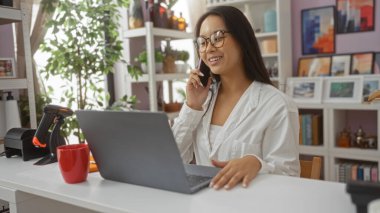 Genç, Asyalı, kadın, ev dekorasyonu mağazasında çalışan, telefonla konuşan ve arka planda bitki ve rafların olduğu dekore edilmiş bir mekanda dizüstü bilgisayar kullanan..