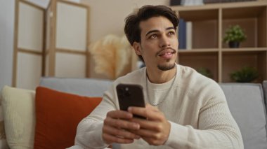 Telefonu olan genç adam, sıcak dekor ve çevre aydınlatması ile çevrili modern bir oturma odasında rahat bir koltukta otururken düşünceli bir şekilde bakıyor ve davetkar bir atmosfer yaratıyor..