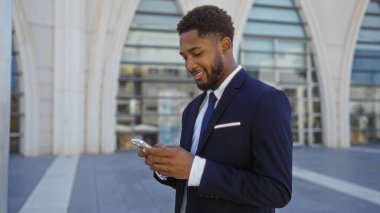Genç, Afrikalı, takım elbiseli Amerikalı adam arka planda modern mimari olan bir şehir sokağında akıllı telefon kullanırken gülümsüyor.