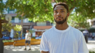 Parlak bir günde yeşil ve arka planda binaları olan bir şehir parkında duran yakışıklı genç adam..