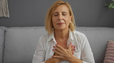 Huzur içinde meditasyon yapan, gözleri kapalı, rahat bir oturma odasında koltukta oturan, elleri kalbinde olan, huzurlu ve mutlu görünen bir kadın.