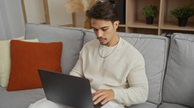 Genç İspanyol adam rahat bir oturma odasında rahat bir koltukta dizüstü bilgisayar kullanıyor modern iç tasarım ve rahat ev atmosferi sergiliyor.