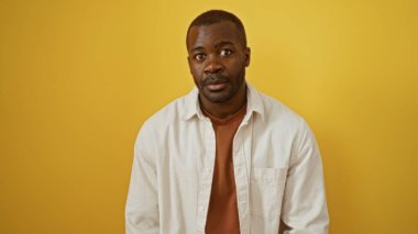 Genç, Afro-Amerikalı bir adam, parlak sarı bir arka planda, günlük beyaz bir gömlekle izole edilmiş.