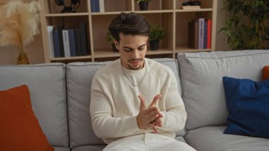 Elleri bir arada olan İspanyol asıllı genç bir adam modern dekorlar ve tarafsız tonlarla çevrili şık bir oturma odasında rahat bir kanepede oturur ve rahat bir ev atmosferi taşır..