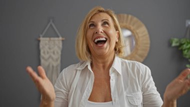 Kısa sarı saçlı, beyaz tişörtlü, orta yaşlı mutlu bir kadın. Keyifli, modern bir oturma odasında, şık dekorlarla gülüyor..