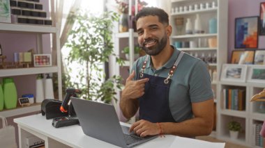 Genç adam dekoratif eşyalarla çevrili bir mağazada çalışırken ve dizüstü bilgisayar kullanırken kendini gösteriyor.
