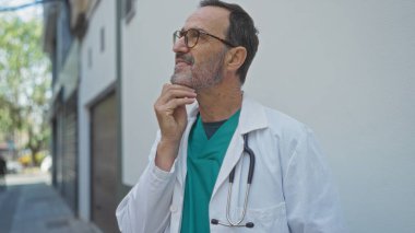 Beyaz önlüklü, steteskoplu, olgun İspanyol bir adam şehir sokağında düşünceli bir şekilde ayakta duruyor..
