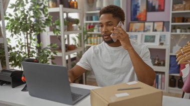 Ev dekorasyon mağazasında dizüstü bilgisayarla çalışan bir adam telefonda konuşuyor. Arka planda bitkiler ve paketler var.