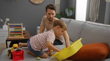 Kanepede oturan bir adam modern evin oturma odasında oyuncak kutusuyla oynayan çocuğu izliyor..