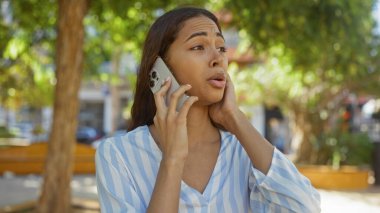 Güneşli bir günde şehir parkında arka planda ağaçlarla telefonda konuşan bir kadın duygularını ifade ediyor..