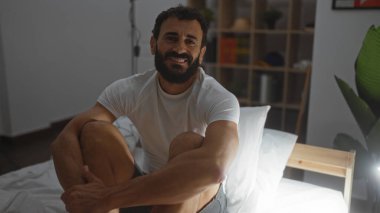 Sakallı genç İspanyol bir adam modern bir iç mekanı olan rahat bir yatak odasında oturuyor..