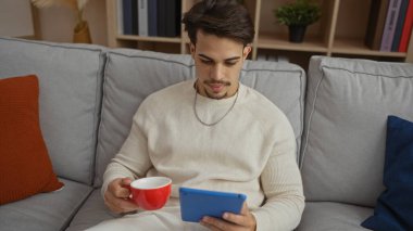 Genç, İspanyol asıllı bir adam, modern bir evde, koltukta tablet ve kahveyle dinleniyor. Konfor, teknoloji ve boş vakitlerini çağdaş bir apartman dairesinde geçiriyor..