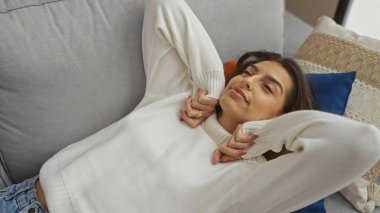 Kapalı koltukta, yastıklarla, beyaz süveter giyip, evinde huzurlu bir şekilde gülümseyen bir kadın.
