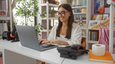 Ev dekorasyonu mağazasında dizüstü bilgisayarı olan bir kadın, masasında sırıtan esmer bir kadın, iç mekan ve dekoratif eşyalarla çevrili, rahat bir iç mekan sergiliyor..