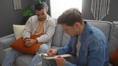 Erkekler rahat bir oturma odasında konuşuyor, biri düşünceli görünüyor, diğeri de bir panoya yazıyor..