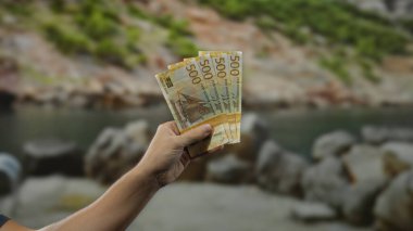 Sahilde kayalar ve su bulunan manzaralı bir sahil bölgesinde Norveçli kronlar tutan bir adam açık havada seyahat ve finans kavramını vurguluyor..