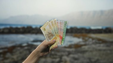 Sahilde deniz manzaralı bir plajda Norveçli banknotlar tutan adam rahat bir açık hava ortamında krone para birimini vurguluyor..