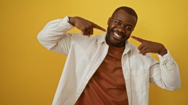 İzole edilmiş sarı bir arka plan duvarında gülümseyen ve kendini işaret eden genç adam, kendine güven ve mutluluk gösteriyor.