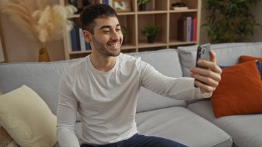 Genç adam rahat oturma odasında selfie çekerken gülümsüyor, modern dekorlar ve rahat mobilyalarla çevrili, evinde sıcak ve davetkar bir atmosfer yaratıyor..