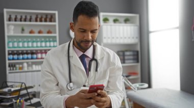 Klinikte raflar ve tıbbi malzemelerle akıllı telefon kullanan İspanyol erkek doktor profesyonel ama rahat bir atmosfer sergiliyor..