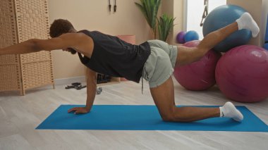 Beden eğitiminde hasırın üzerinde egzersiz yapan bir adam içeride, spor aletleri görünür vaziyetteyken, şort ve kolsuz tişört giyerken poz veriyor.